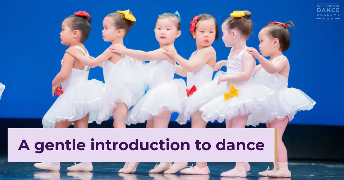 Children in white tutus dancing in line on stage with colored ribbons.